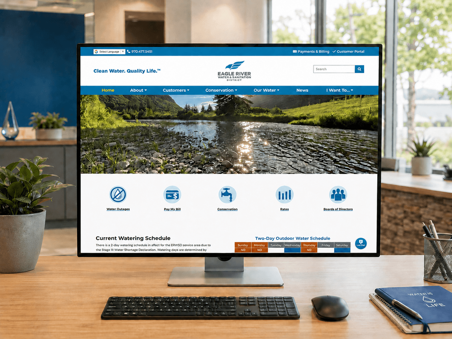 Eagle River Water & Sanitation District website displayed on a desktop monitor in a modern office workspace with plants, a keyboard, mouse, and water-themed desk accessories.